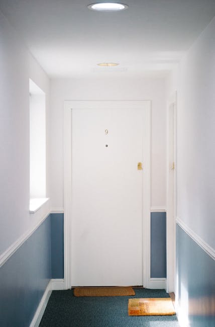 A clean, well-lit hallway featuring a white door with the number 9, positioned centrally, with a small peephole and a brass-colored handle. The walls are painted white on the upper half and a muted bl