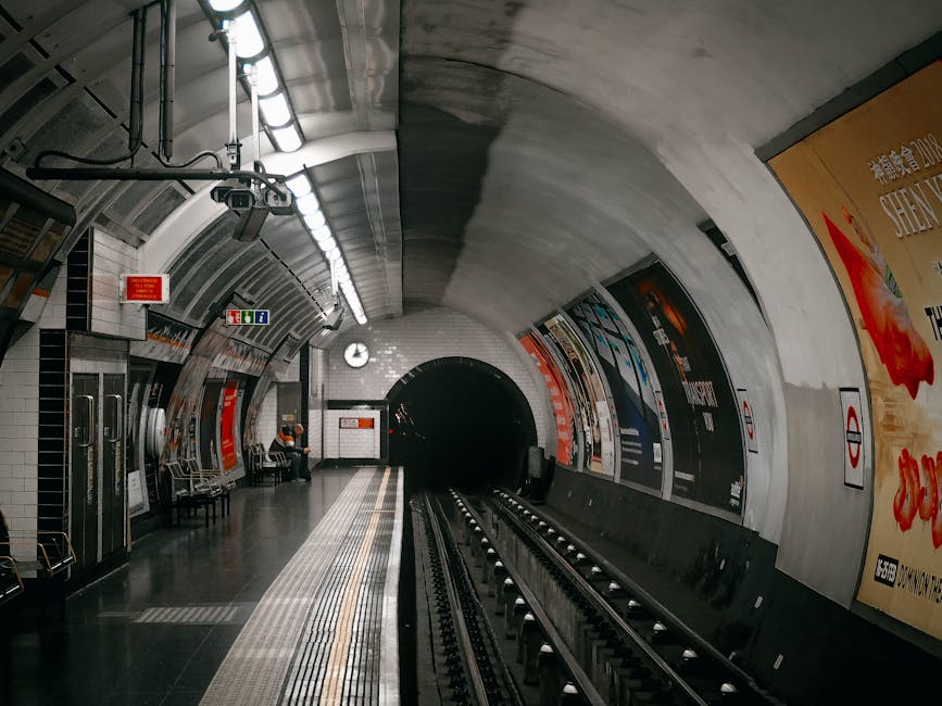 A spacious underground subway station corridor with tiled floors and curved white ceiling panels illuminated by linear lighting. On the left, the tiled wall features the London Underground roundel sig